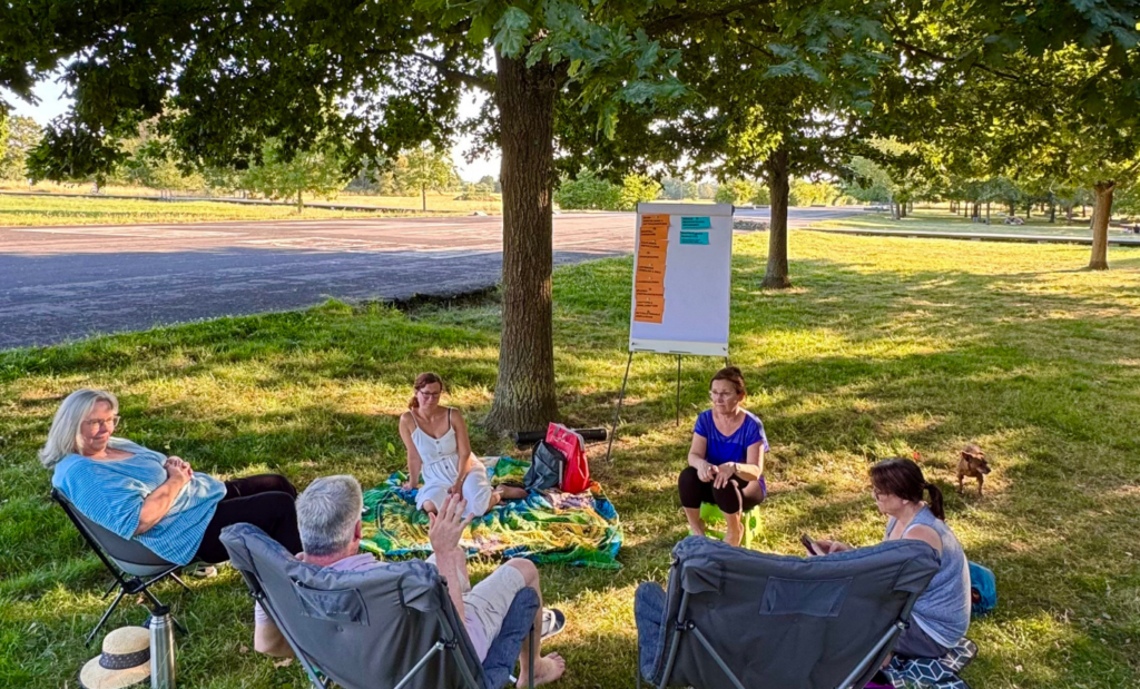 Kleine Gruppe von Erwachsenen sitzt im Freien unter Bäumen im Kreis bei einer Infoveranstaltung mit Flipchart.