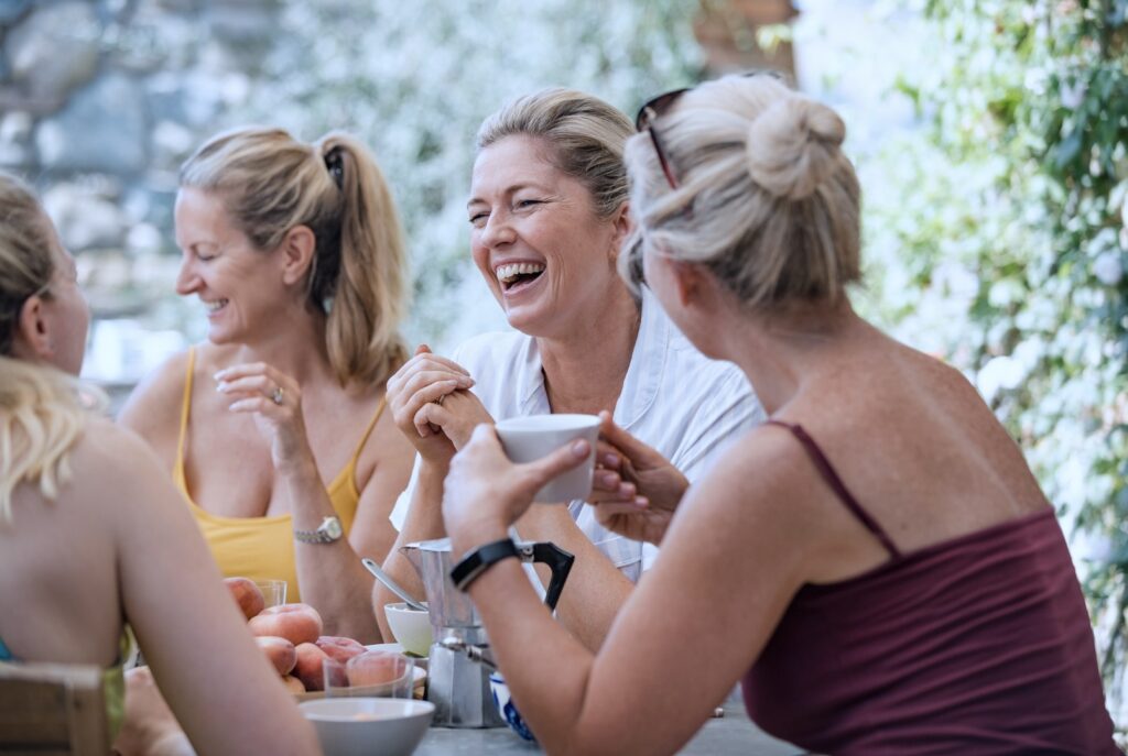 Vier Frauen lachen bei einem Treffen im Freien und trinken gemeinsam Kaffee, Symbol für Austausch und Lebensqualität.